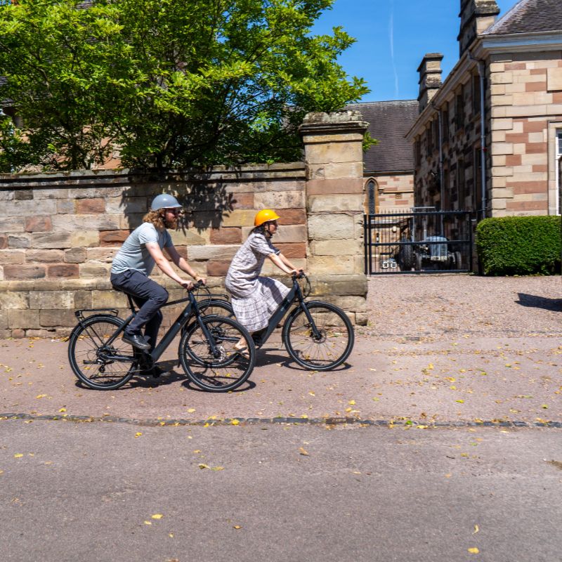 Dawes Scenic Crossbar ebike lifestyle Two people riding on a path in front of a stone building with trees in the background.