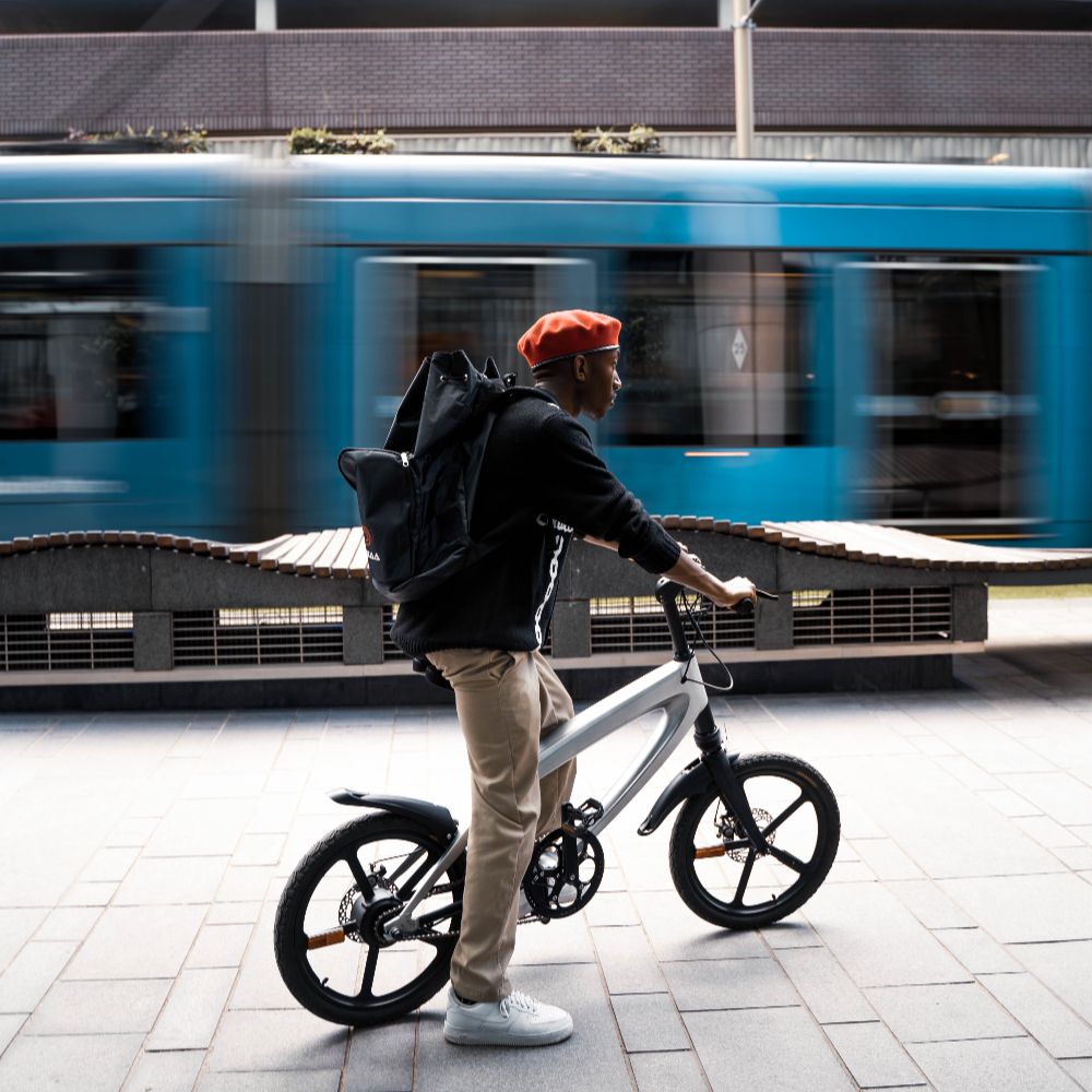 Cruzaa City e-bike lifestyle image of a Person with a backpack standing on a sidewalk with a blurred blue train in the background