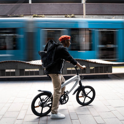 Cruzaa City e-bike lifestyle image of a Person with a backpack standing on a sidewalk with a blurred blue train in the background