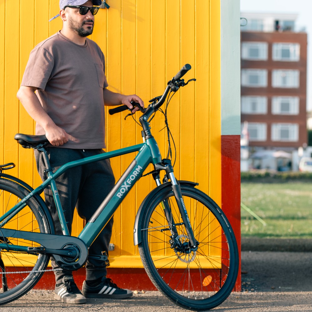 Roxform Valor S e-bike lifestyle image Man standing next to the bicycle against a yellow wall with a building in the background